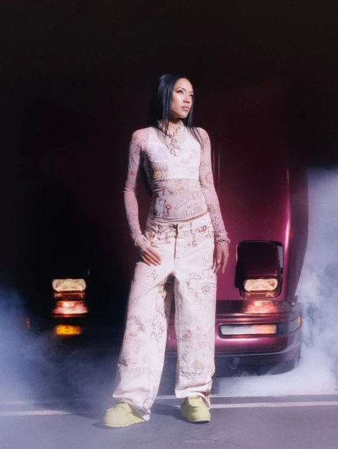 Woman in patterned top and pants poses in front of burgundy car with haze effect. Fashion shot, studio setting, diffused lighting.