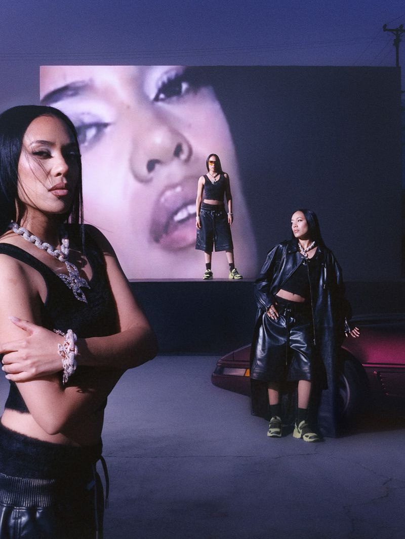 Three women in fashionable outfits pose in an outdoor setting. A large screen shows a close-up of a woman's face in the background.