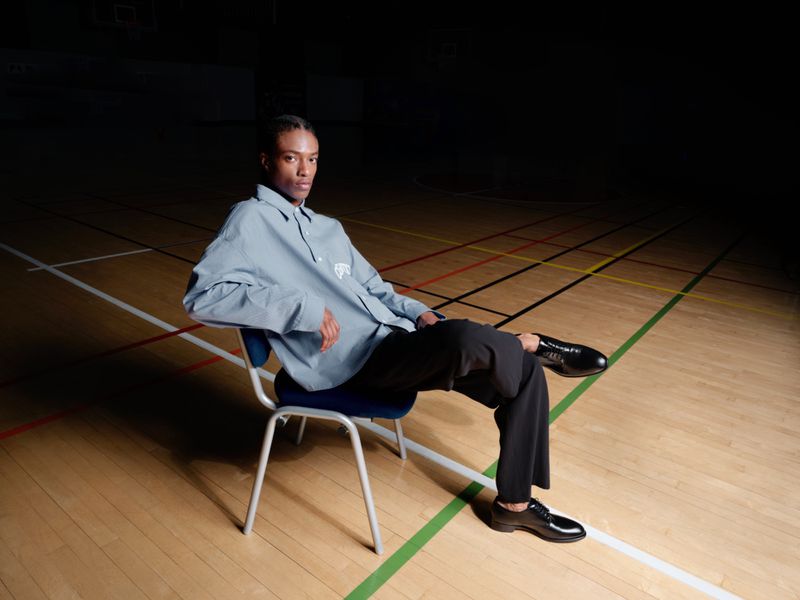 A stylish, slender Black male model sits on a blue chair in a gymnasium, wearing a blue button-down shirt and black dress pants and shoes.