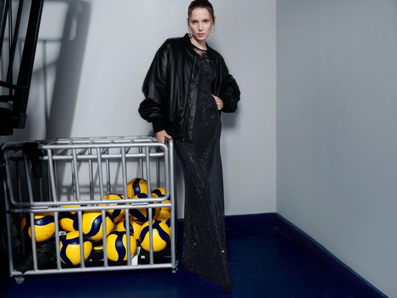 A woman in an evening gown and bomber jacket poses next to a metal bin full of volleyballs in a studio with blue and gray walls.