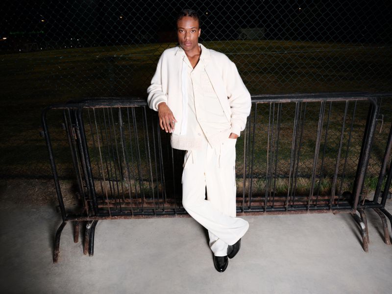 A solitary figure in fashionable attire leans against a metal railing in an outdoor setting under the soft glow of evening lights.