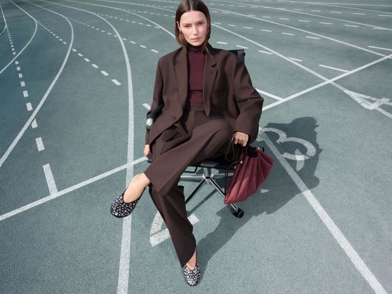 Woman in brown suit sits on office chair on running track with red bag, wearing studded ballet flats and burgundy top, looking at the camera.