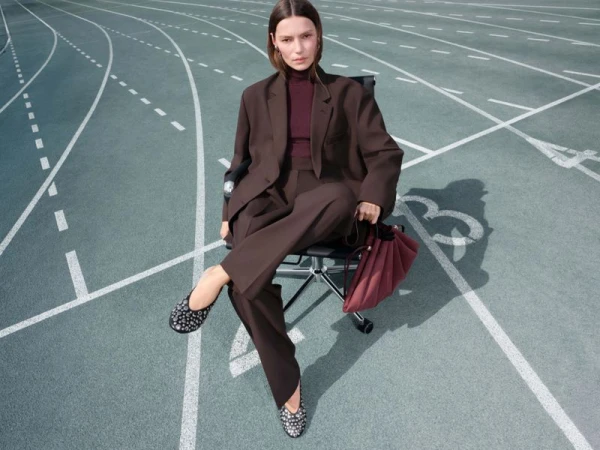 Woman in brown suit sits on office chair on running track with red bag, wearing studded ballet flats and burgundy top, looking at the camera.