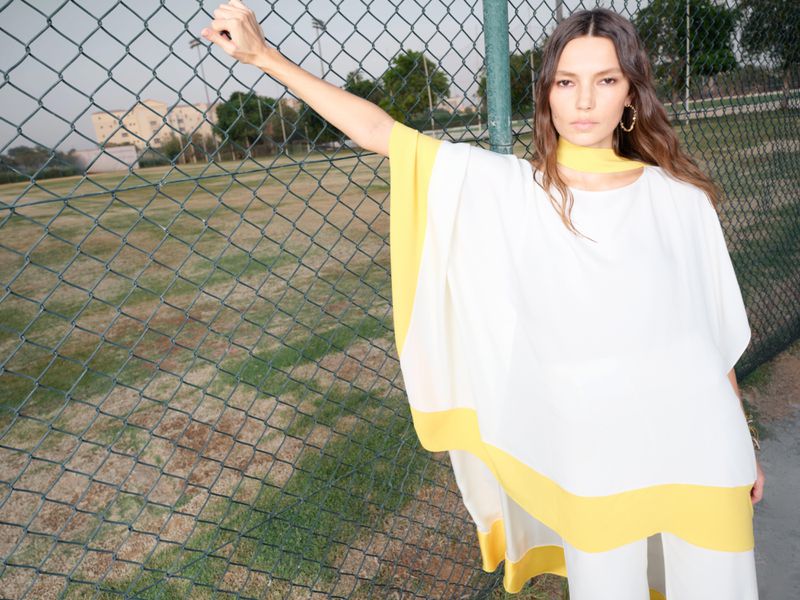 Woman in a white and yellow poncho stands by a chain-link fence in an outdoor setting, showcasing contemporary fashion apparel.