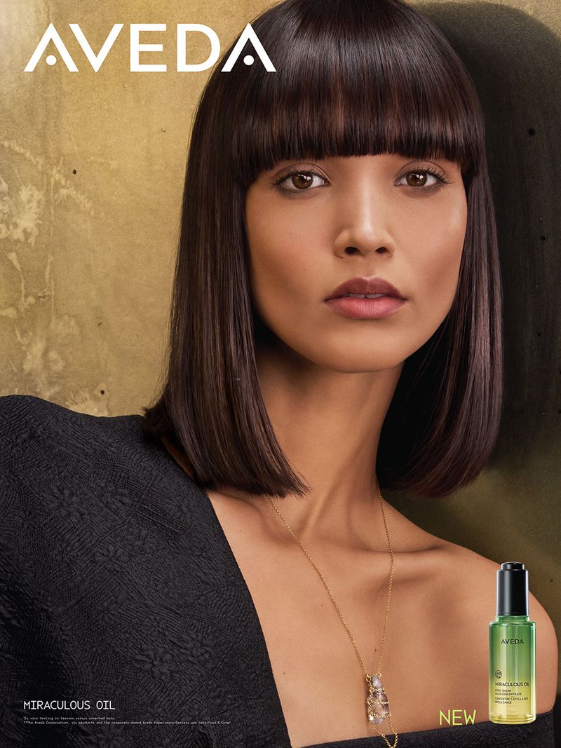 Portrait of a woman with dark hair and bangs, wearing a black top and gold necklace, next to a bottle of hair oil.