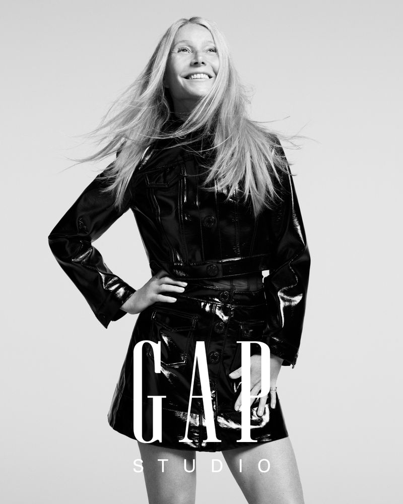 Smiling adult female with long blonde hair posing in shiny black Gap brand jacket and skirt in front of white studio background.