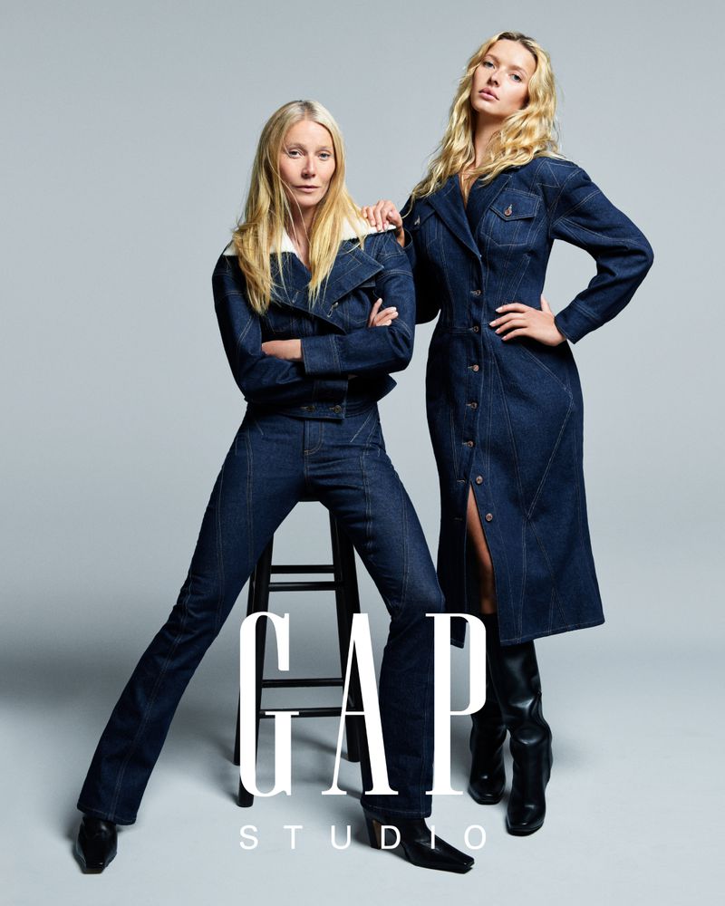 Two women in denim outfits pose in a studio shot for a GAP ad campaign, featuring a stool, a white backdrop, and the GAP logo.