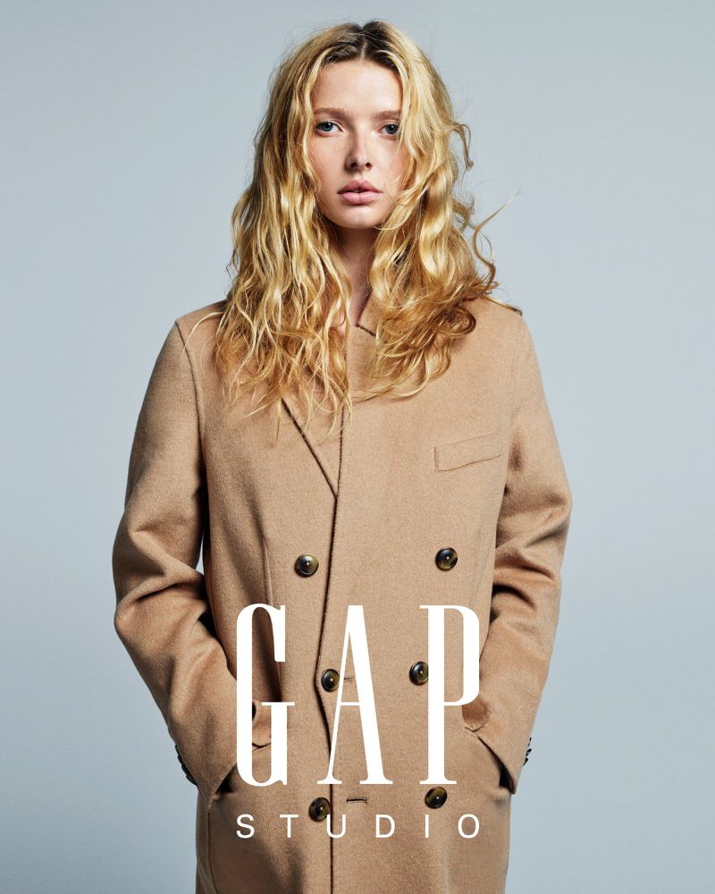 Fashion portrait of a young fair-skinned woman with blonde wavy hair wearing a tan wool double-breasted overcoat in a photo studio.