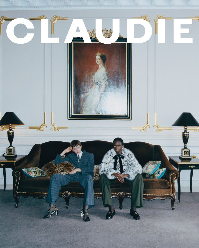 Two fashion models pose on a vintage brown couch in an indoor setting. One wears a blue suit, the other a faux fur coat and green pants.