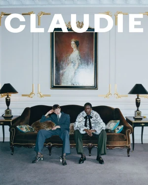 Two models in tailored and fur coats sitting on an antique brown sofa beneath a framed portrait in an ornate room.