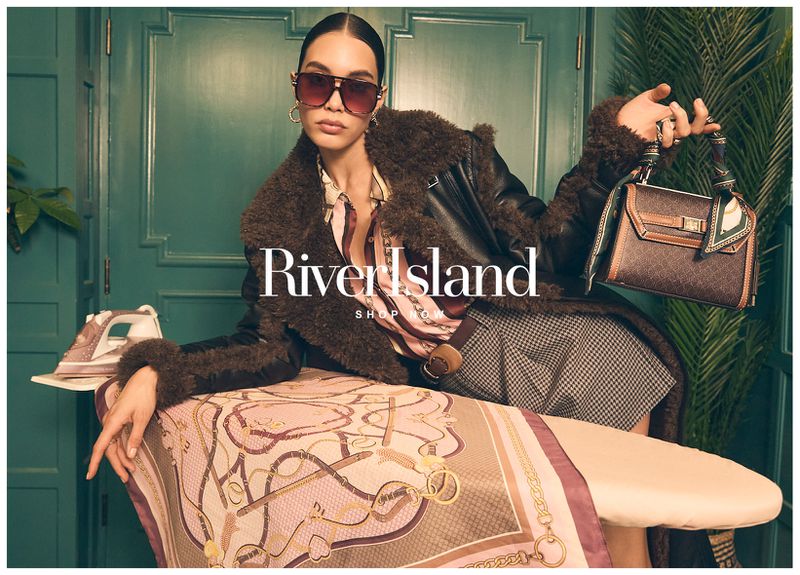 Fashion editorial shot featuring an adult woman wearing sunglasses and a brown jacket, posing with a handbag in an indoor setting.