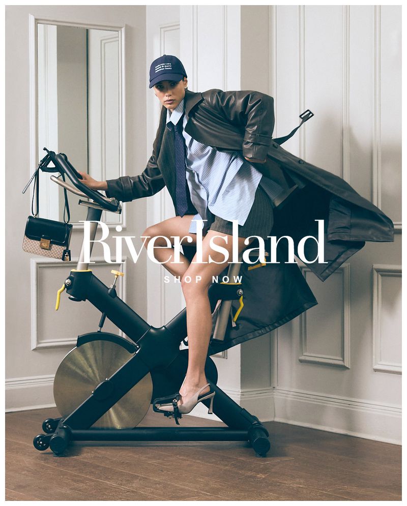 Fashion-forward woman in a trench coat exercises on a stationary bike, accessorized with a handbag and baseball cap in a staged indoor studio.