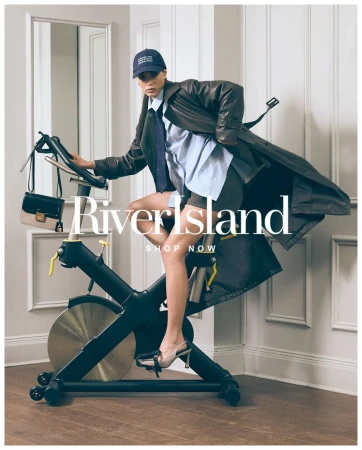 Fashion-forward woman in a trench coat exercises on a stationary bike, accessorized with a handbag and baseball cap in a staged indoor studio.