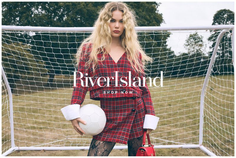 Blonde model in red plaid jacket and skirt, holding a soccer ball, standing in front of a soccer net on a grassy field.