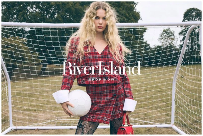 Blonde model in red plaid jacket and skirt, holding a soccer ball, standing in front of a soccer net on a grassy field.