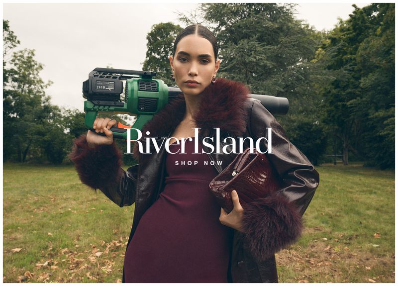Fashion model poses outdoors wearing a dark coat with fur trim, holding a purse, and carrying a green electronic device on her shoulder.