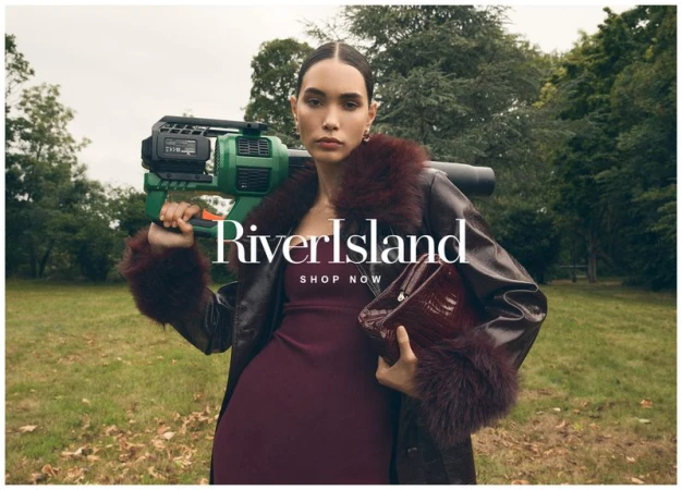 Fashion model poses outdoors wearing a dark coat with fur trim, holding a purse, and carrying a green electronic device on her shoulder.