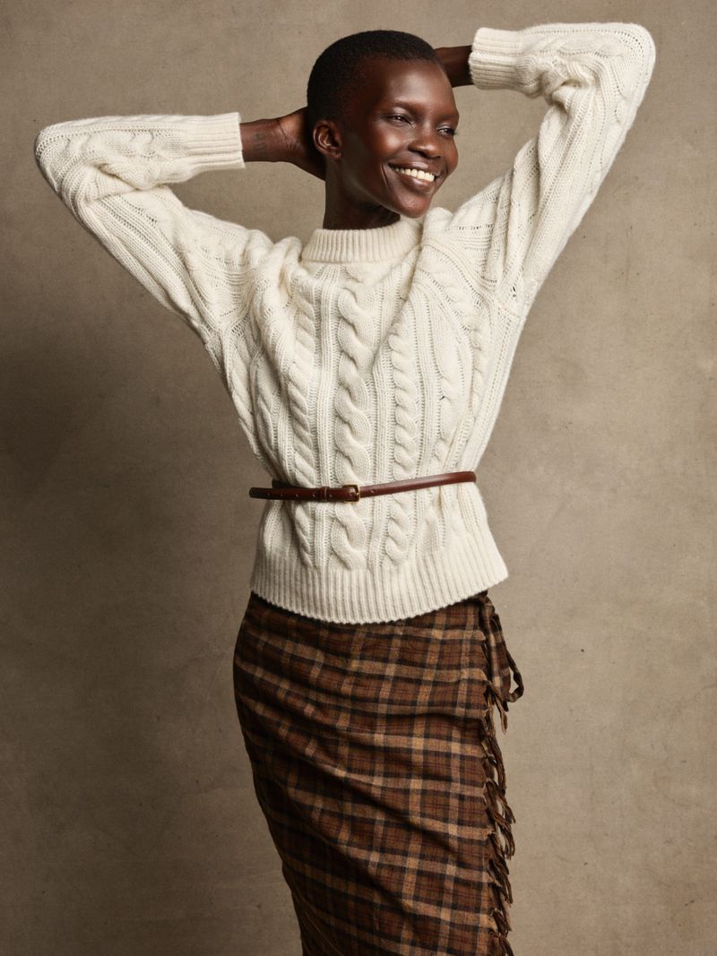 Woman in a cream cable-knit sweater and plaid skirt with hands behind her head, smiling and looking off to the side. Fashion shot.