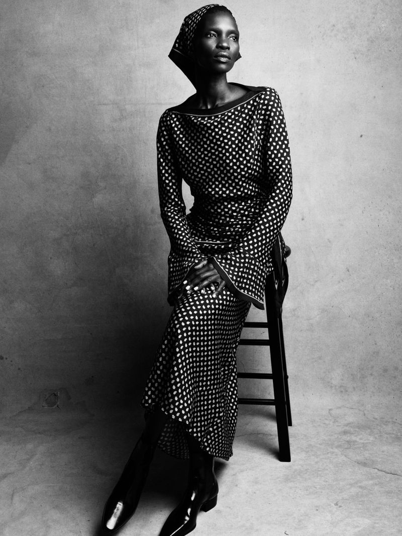 Model Achok seated on a dark wooden chair wearing a patterned dress and matching headwrap in a studio setting.