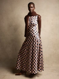 Model in plaid maxi dress and brown scarf standing against a textured studio background for the DÔEN Fall 2025 campaign.