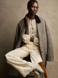 Model Achok wearing a cream lace top, silk trousers with lace hem, and a tweed coat over her shoulders against a textured beige backdrop.
