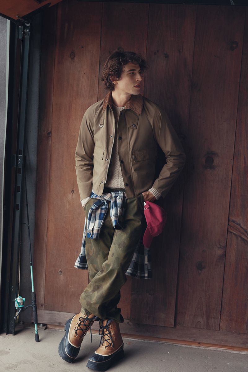 A young male model poses against a wooden wall, wearing a khaki jacket, patterned sweater, camouflage pants, and boots, holding a red cap.