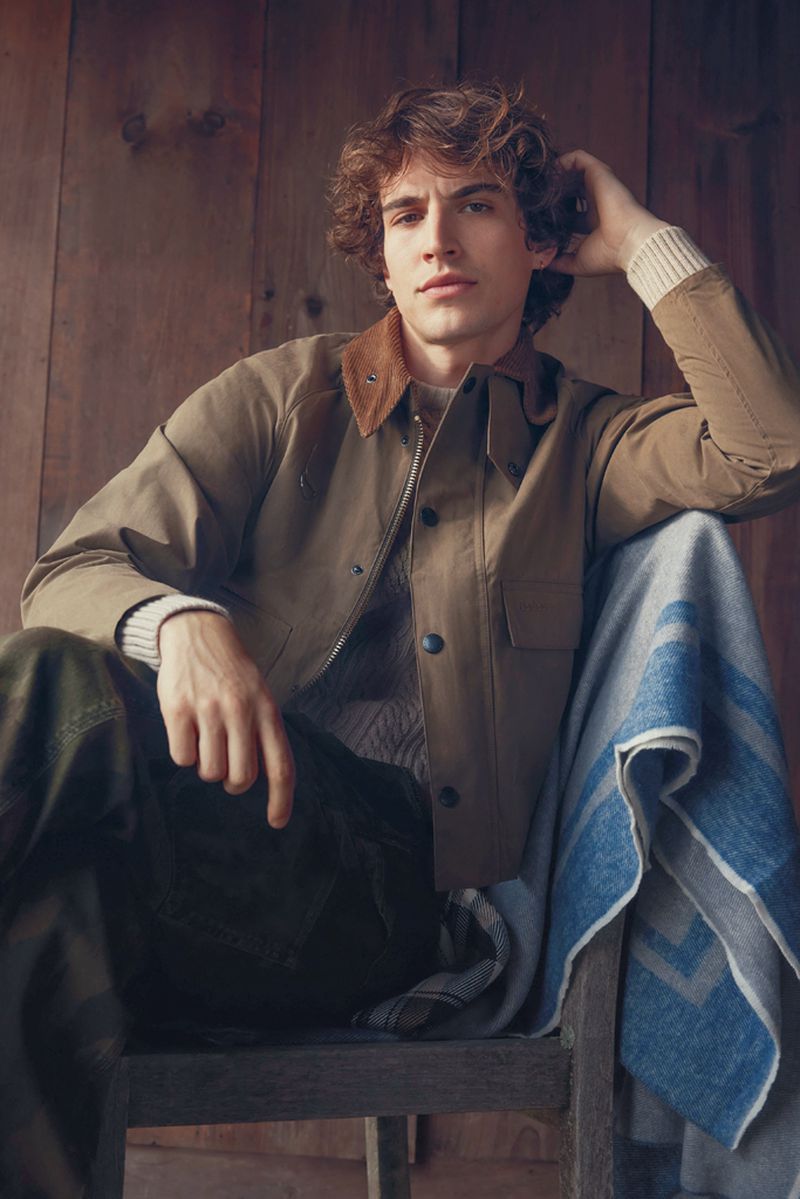 Young man with curly brown hair wearing a tan jacket and sweater seated against a rustic wood wall