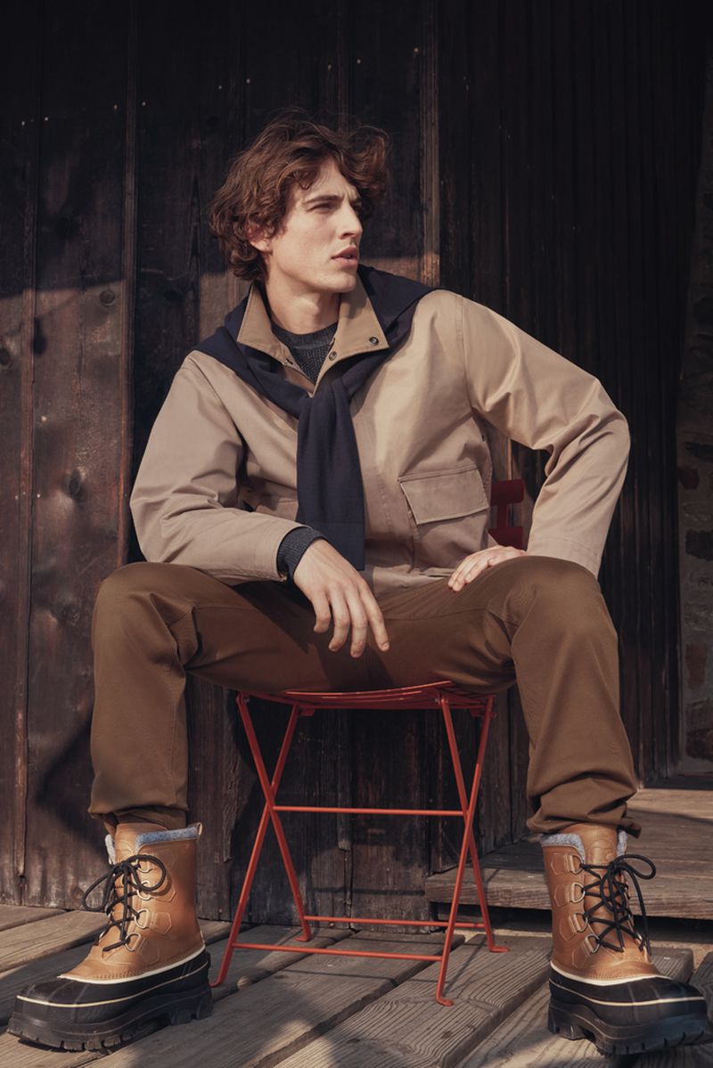 Male fashion model in a jacket, sweater, and boots seated on a red chair outdoors against a weathered wood backdrop, casual and stylish.