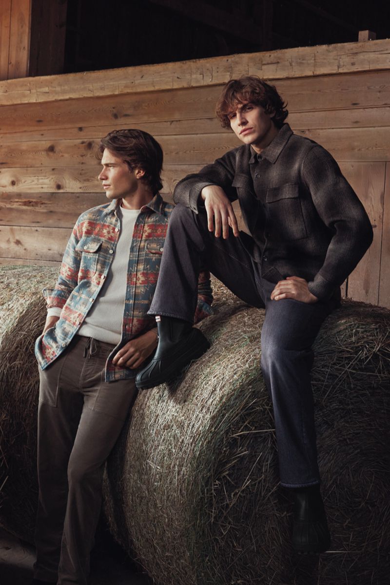 Two men in casual fashion attire pose in a rustic barn setting, leaning against hay bales, showcasing comfortable and stylish outdoor wear.