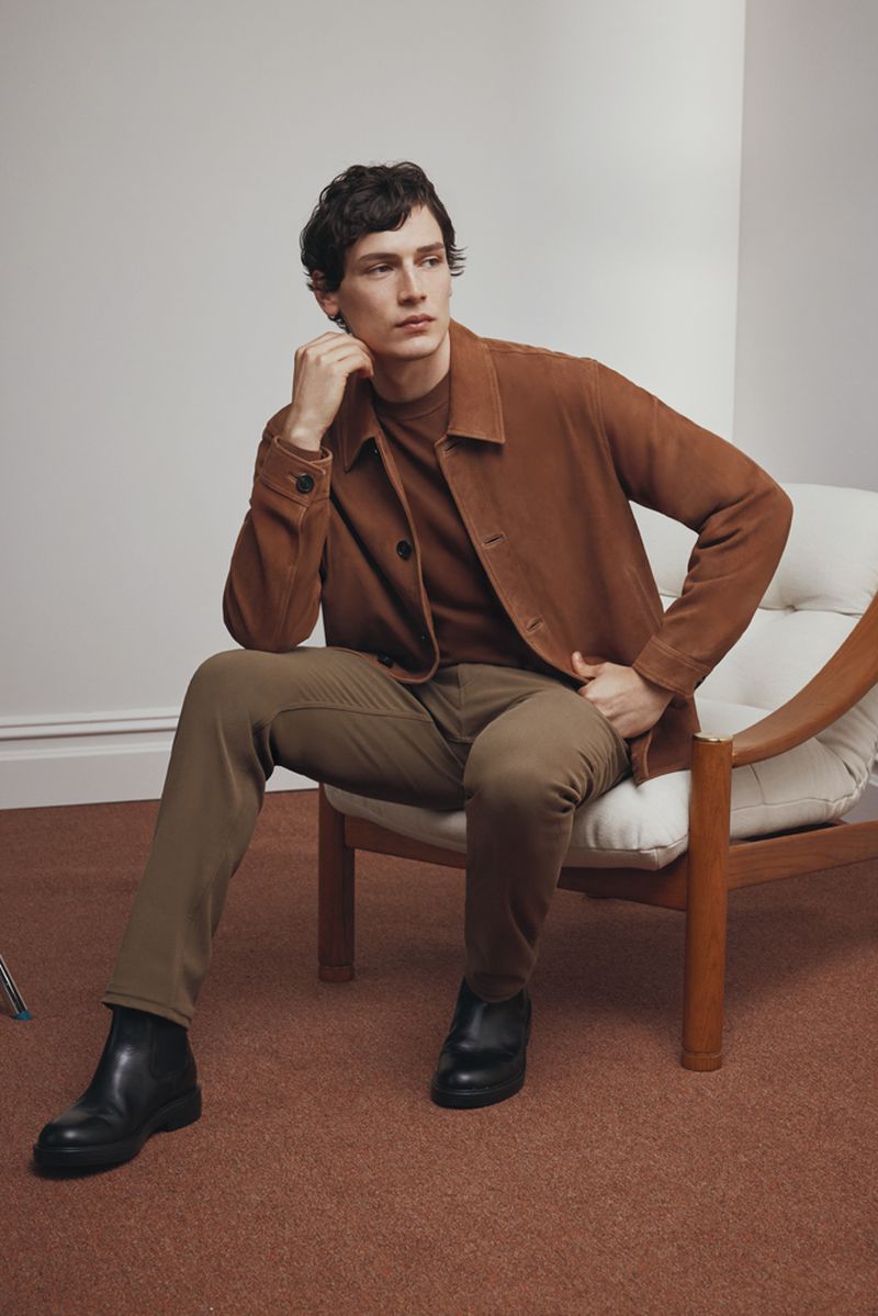 A man wearing a brown jacket and trousers sits in a chair against a neutral background, showcasing modern casual fashion.