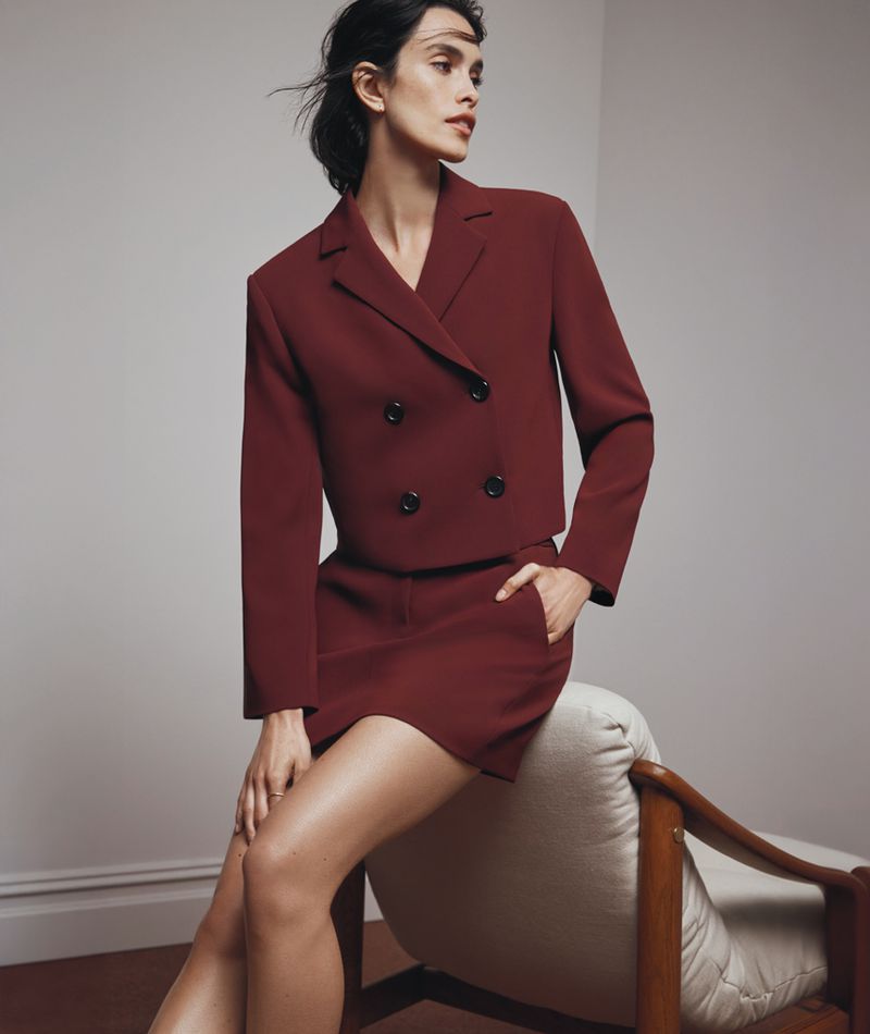 A brunette woman in a burgundy suit is seated on a chair in a studio setting, showcasing a sophisticated and stylish fashion look.