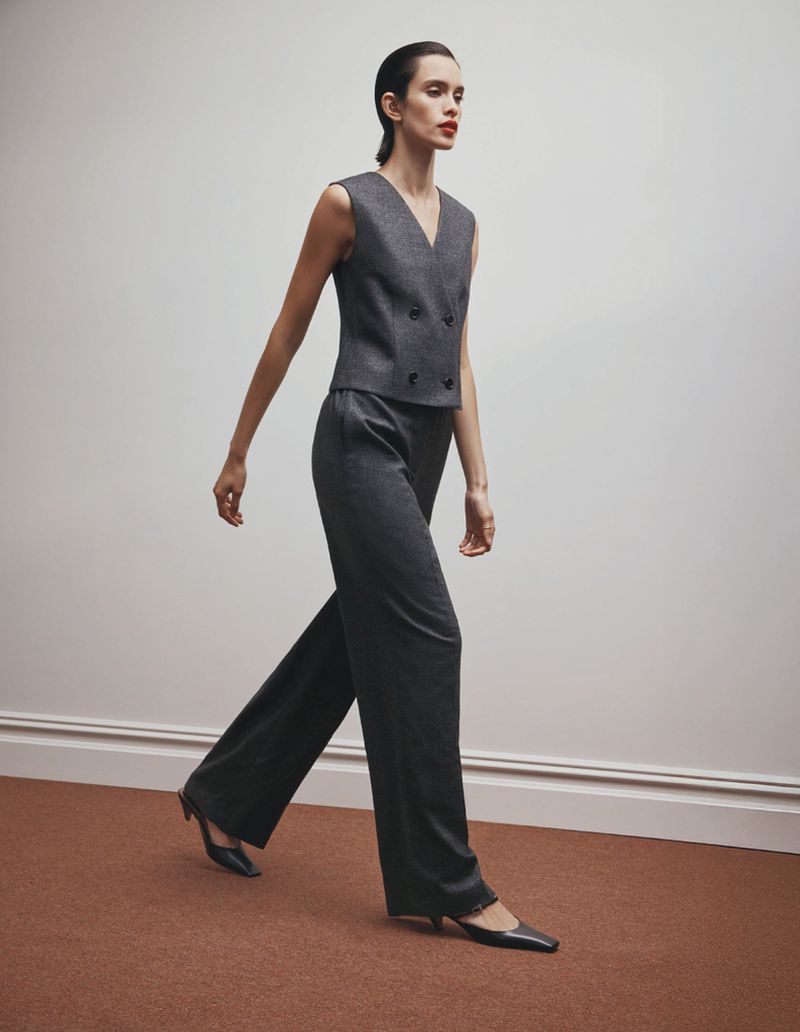 A slender woman in a sleeveless grey suit walks on a brown carpet against a white wall, showcasing minimalist fashion and sleek design.