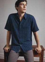 Young man wearing a navy blue pinstripe shirt seated and leaning on a dark wooden table against a plain light grey wall.