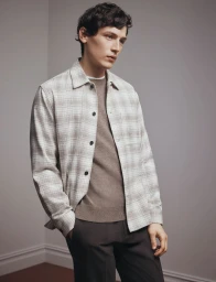 A male model wearing an unbuttoned light plaid overshirt over a taupe knit sweater and dark trousers against a neutral wall.