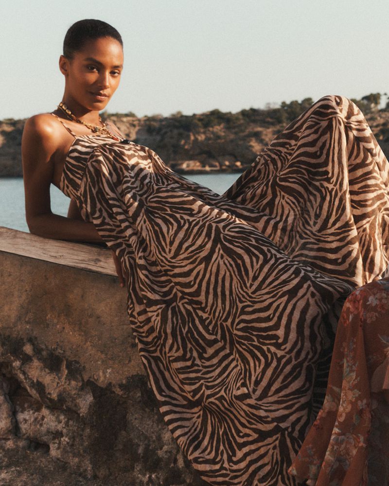 A woman in a long flowing dress with a zebra print poses outdoors near the sea with rocks and low vegetation in the background on a sunny day.