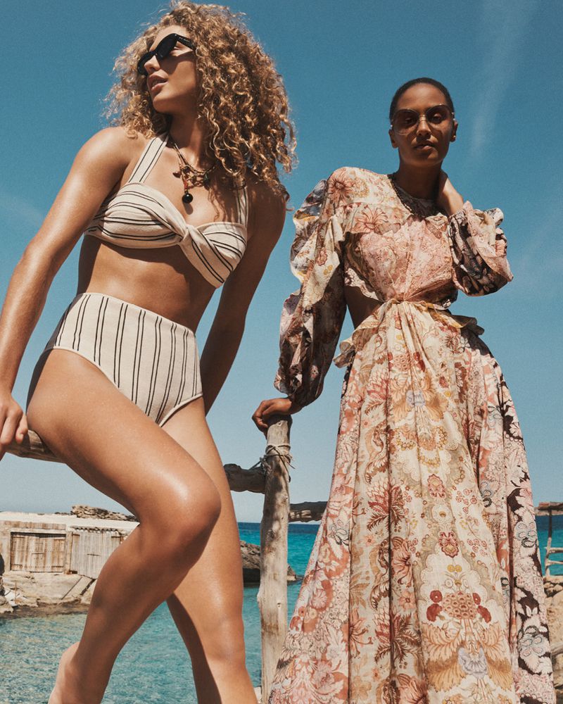 Two women pose in swimwear and a dress against a backdrop of blue sky and turquoise sea. One wears a striped bikini; the other, a floral dress.