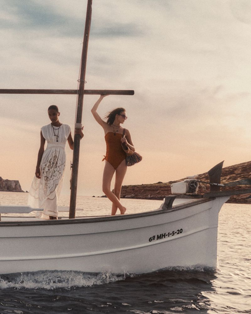 Two women in swimwear and cover-up pose on a small boat at sea against a sunset sky with coastal land in the background.