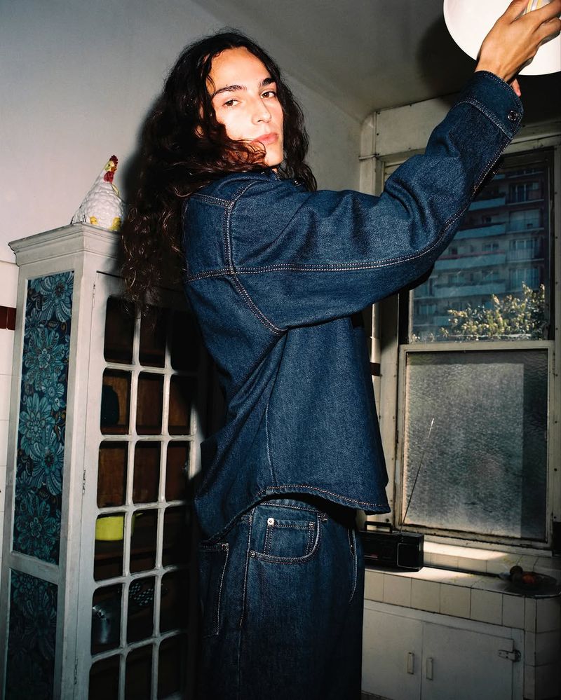 Young adult wearing a denim jacket in a kitchen reaches up toward a ceiling light, showing an interior scene with natural lighting.