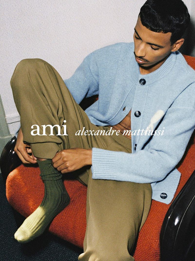 Man wearing a light blue cardigan and olive trousers sitting in an orange chair adjusting his sock