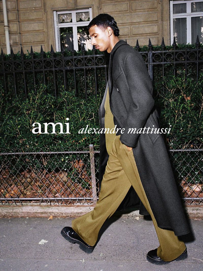 Man in a dark coat and olive trousers walks on a city sidewalk, showcasing a stylish urban look with AMI branding in the background.