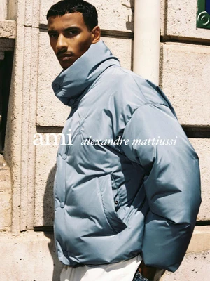 Male model wearing a light blue puffer jacket with a high collar standing against an exterior stone wall.