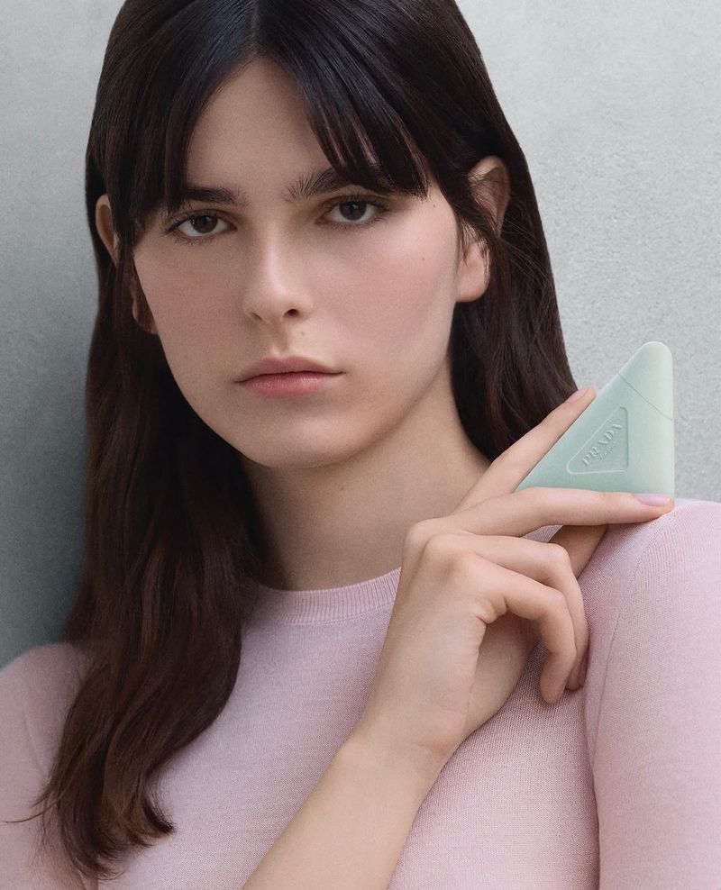 Portrait of a brunette woman holding a triangular, pale-green Prada object. She wears a light-pink sweater against a grey background.