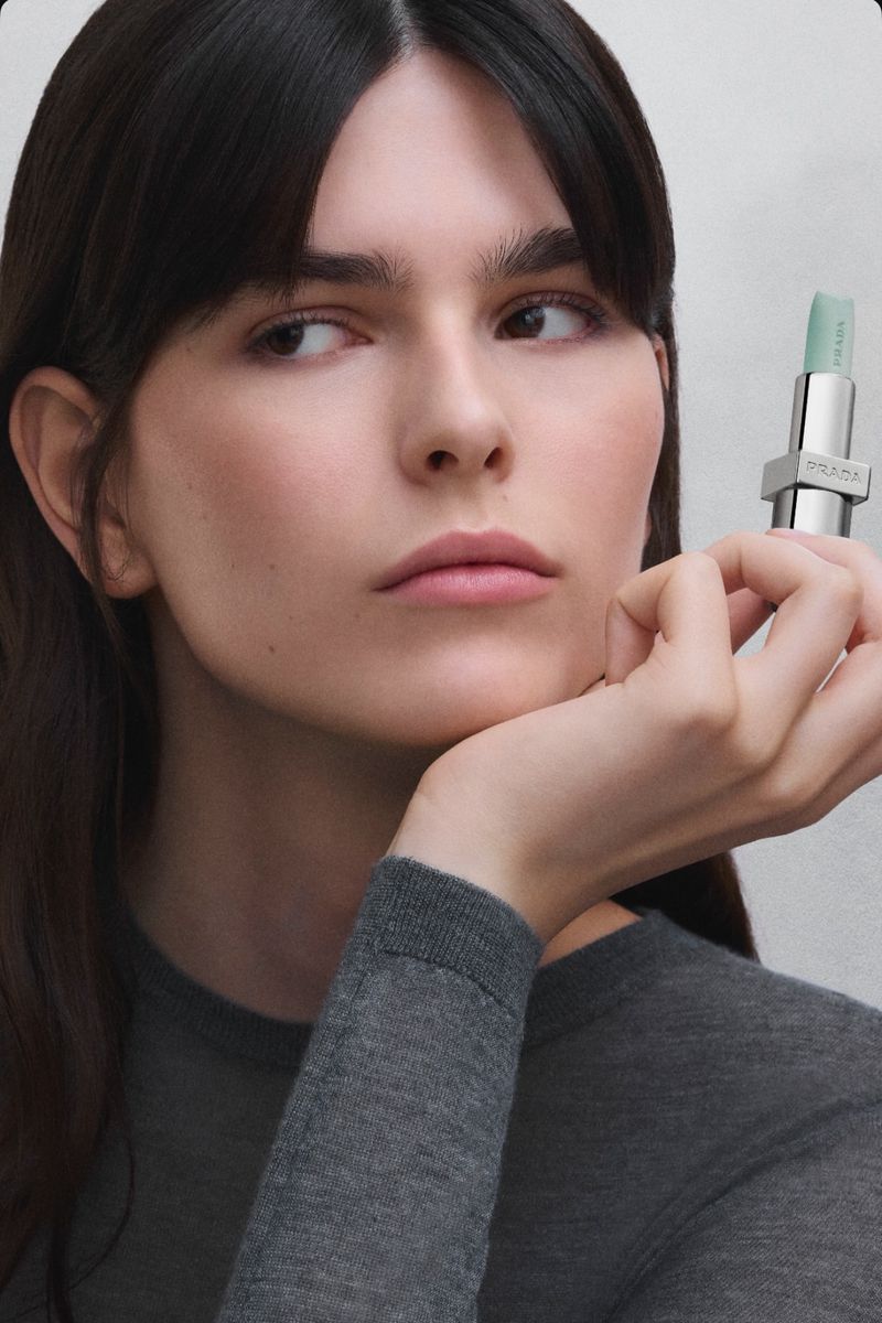 Close-up of a woman with dark hair holding a mint green lipstick. She wears a gray sweater, set against a neutral background.