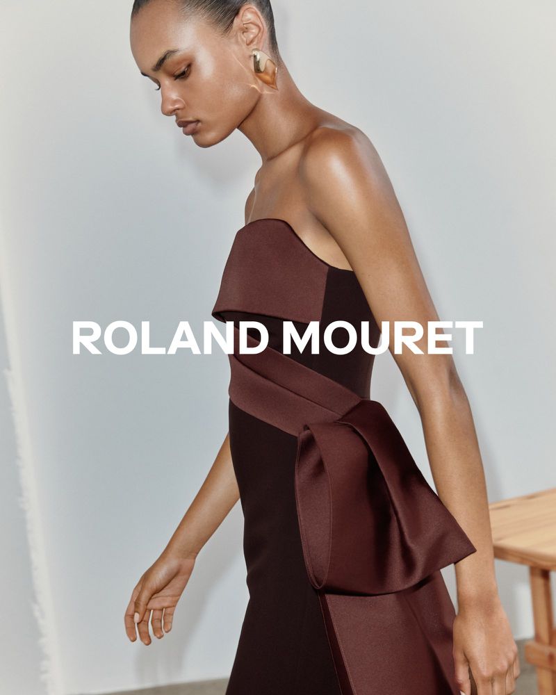 Fashion model in a strapless brown Roland Mouret dress with sculptural bow detail. Studio shot, neutral background, simple wood prop visible.