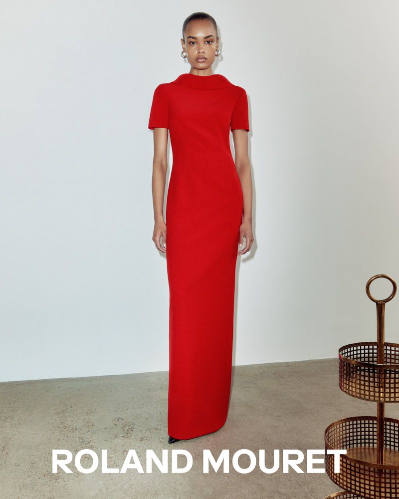Elegant adult female in a long red dress standing in front of a white wall in a studio setting, showcasing high fashion.