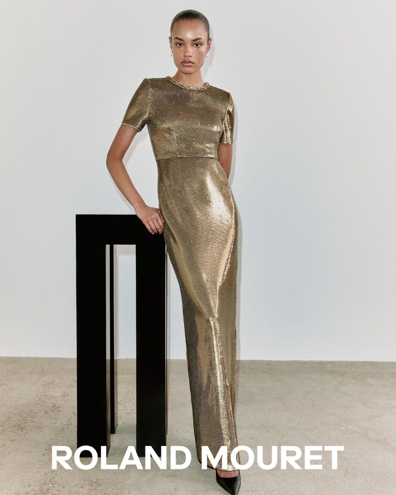 Fashion model in a gold metallic Roland Mouret dress posing in a studio with a black geometric pedestal, minimalist aesthetic.