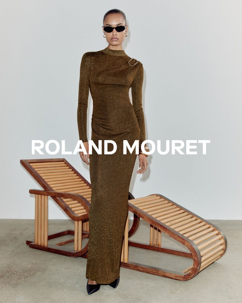 A woman poses in a long-sleeved gold dress with black sunglasses in a studio with natural wood furniture and a clean, minimalist aesthetic.