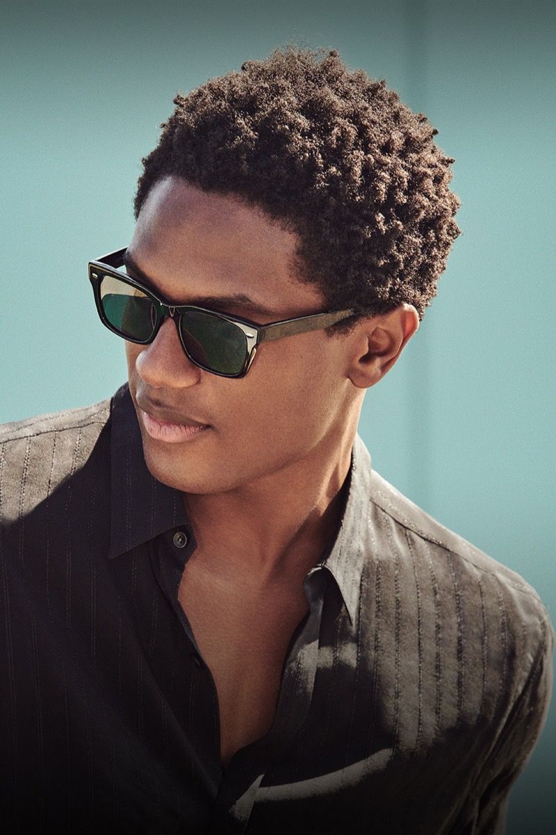 Model Hamid Onifade wearing black sunglasses and an open dark button-down shirt against a soft muted teal background.