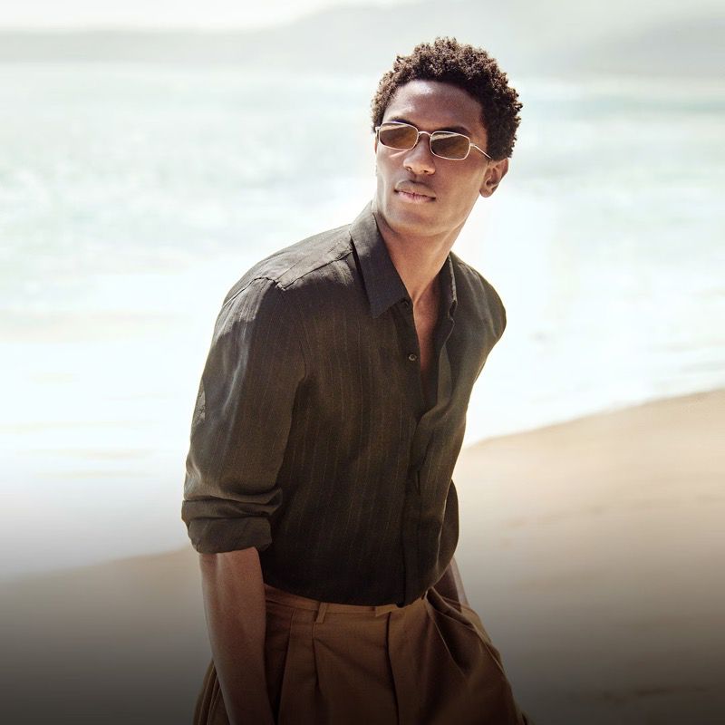 A dark-haired man in a linen shirt and sunglasses poses on a beach with a soft focus and natural light in this fashion shot.