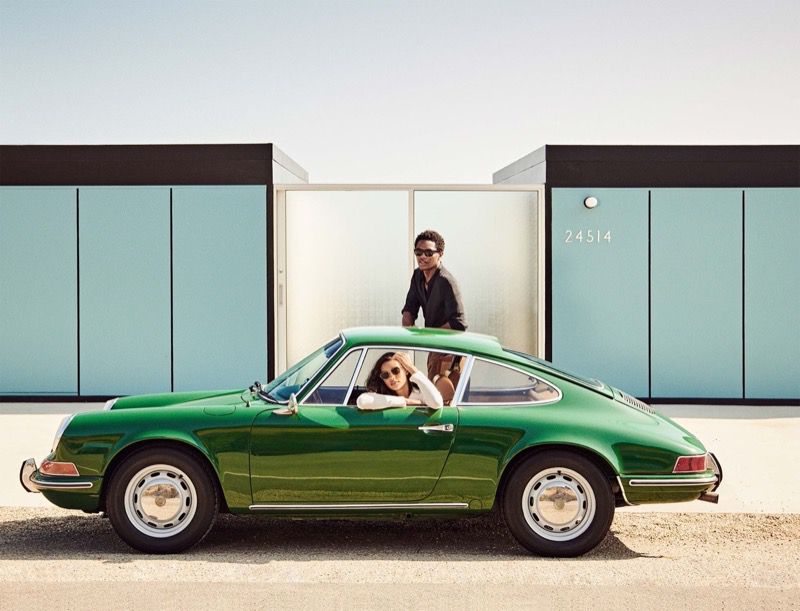 Man and woman wearing sunglasses posing with a vintage green sports car parked in front of modern architecture.